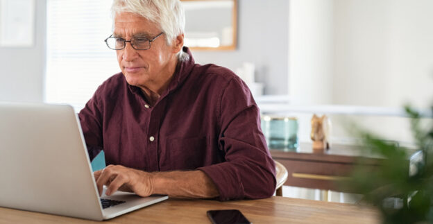 Grey headed man using a laptop on table at home.
